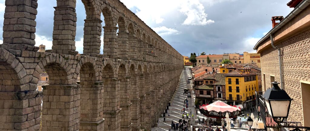 View of the Roman Aqueduct in Segovia, featuring its iconic arches and surrounding buildings under a partly cloudy sky.