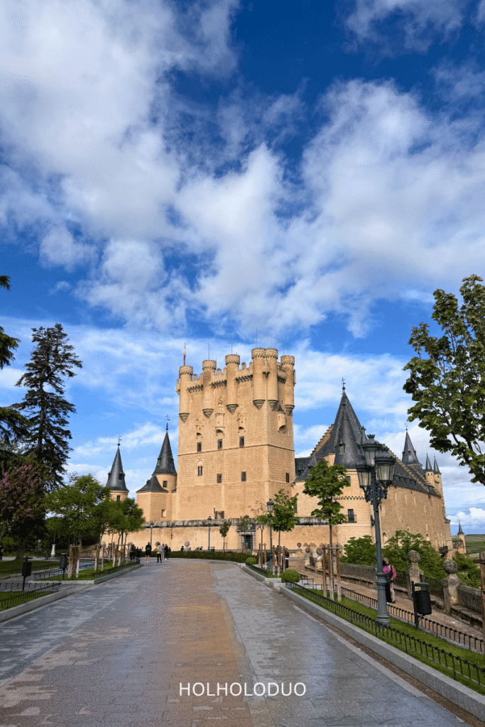 A scenic view of the Alcázar of Segovia, showcasing its fairy-tale-like towers and turrets against a vibrant blue sky and scattered clouds, with a pathway leading up to the castle flanked by trees.